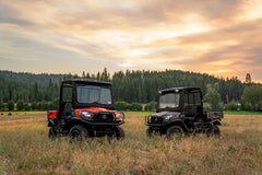Two Kubota RTV-X utility vehicles, orange and camouflage, with roofs and windshields, in a meadow at sunset