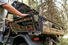 A hand lowering the tailgate of a camouflage Kubota RTV-X utility vehicle's cargo box