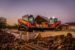 Two Kubota SVL series compact track loaders with buckets on a building site at dusk, their work lights on