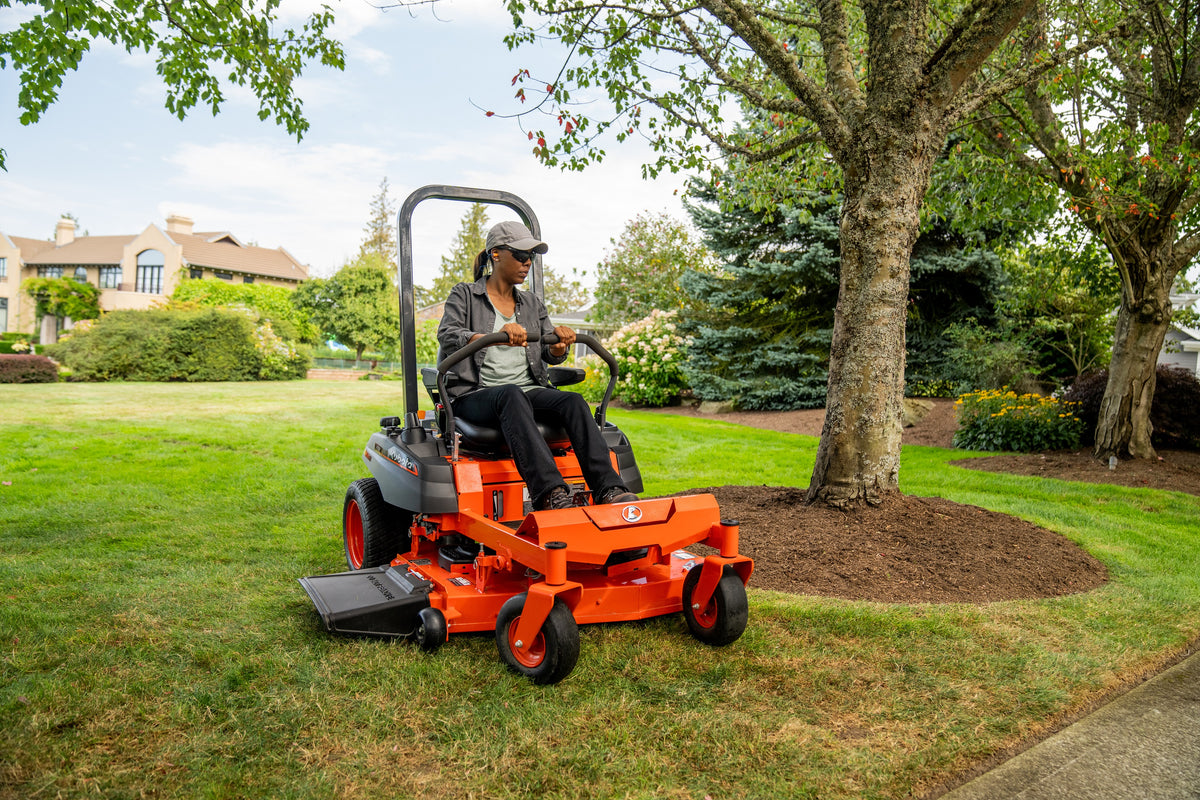 Kubota Z232 zero-turn mower mowing beside a tree on a large lawn, a house in the distance