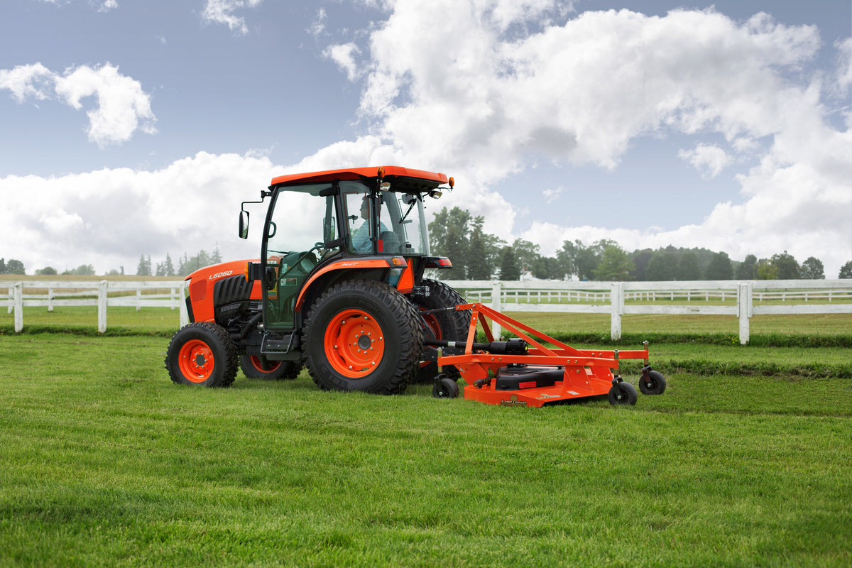 Kubota tractor with a cab pulling a Land Pride FDR25 series grooming mower across a lawn by a white fence