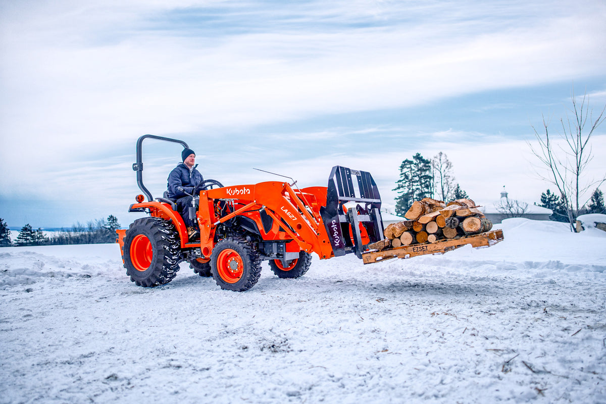 Kubota L series tractor with an LA627 front loader, carrying firewood on pallet forks in the snow