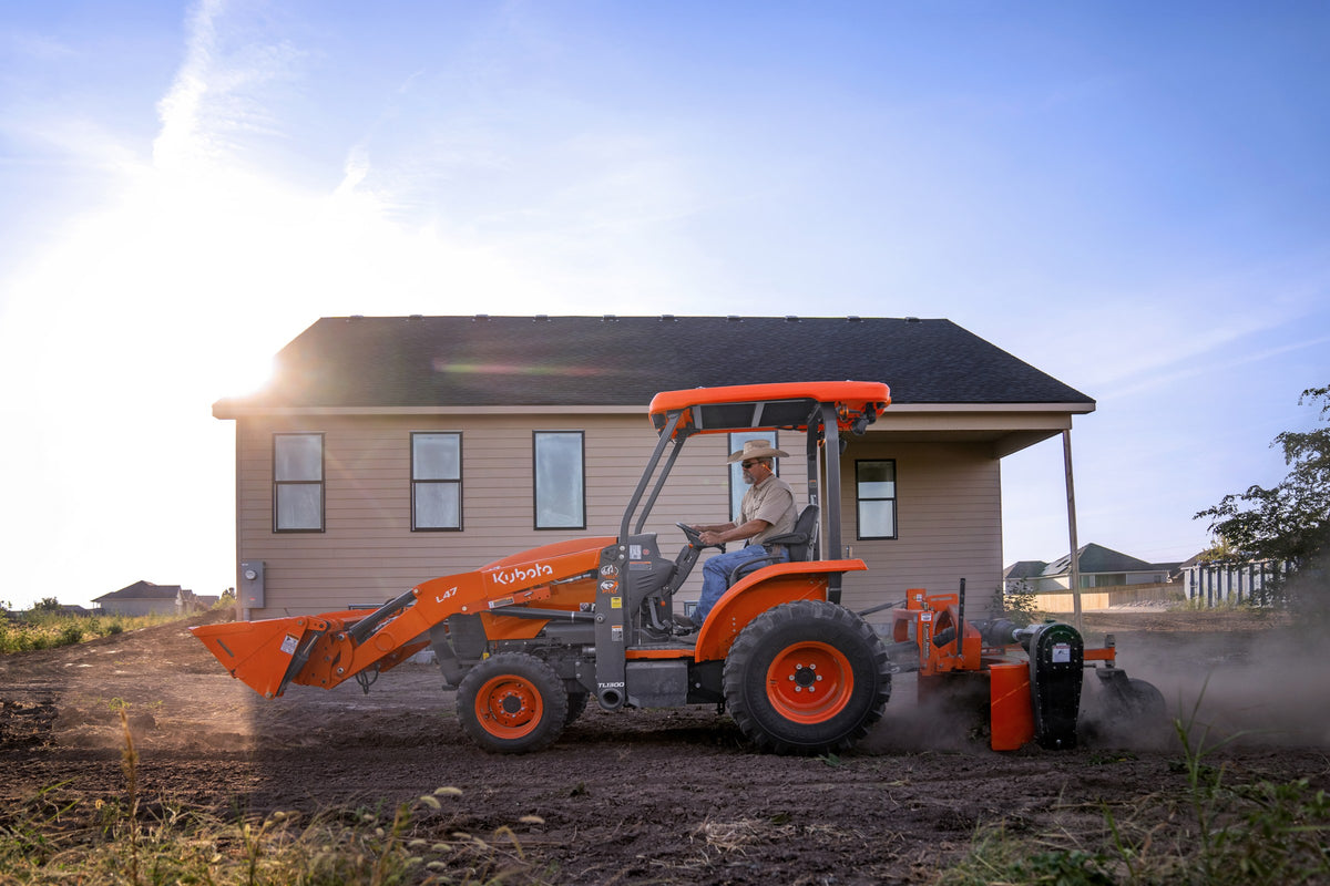 Kubota L47 tractor-loader-backhoe working soil beside a new house, a rotary tiller on the back