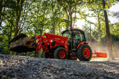 Kubota L6070 ProCab carrying logs in a loader grapple on a gravel road through the woods