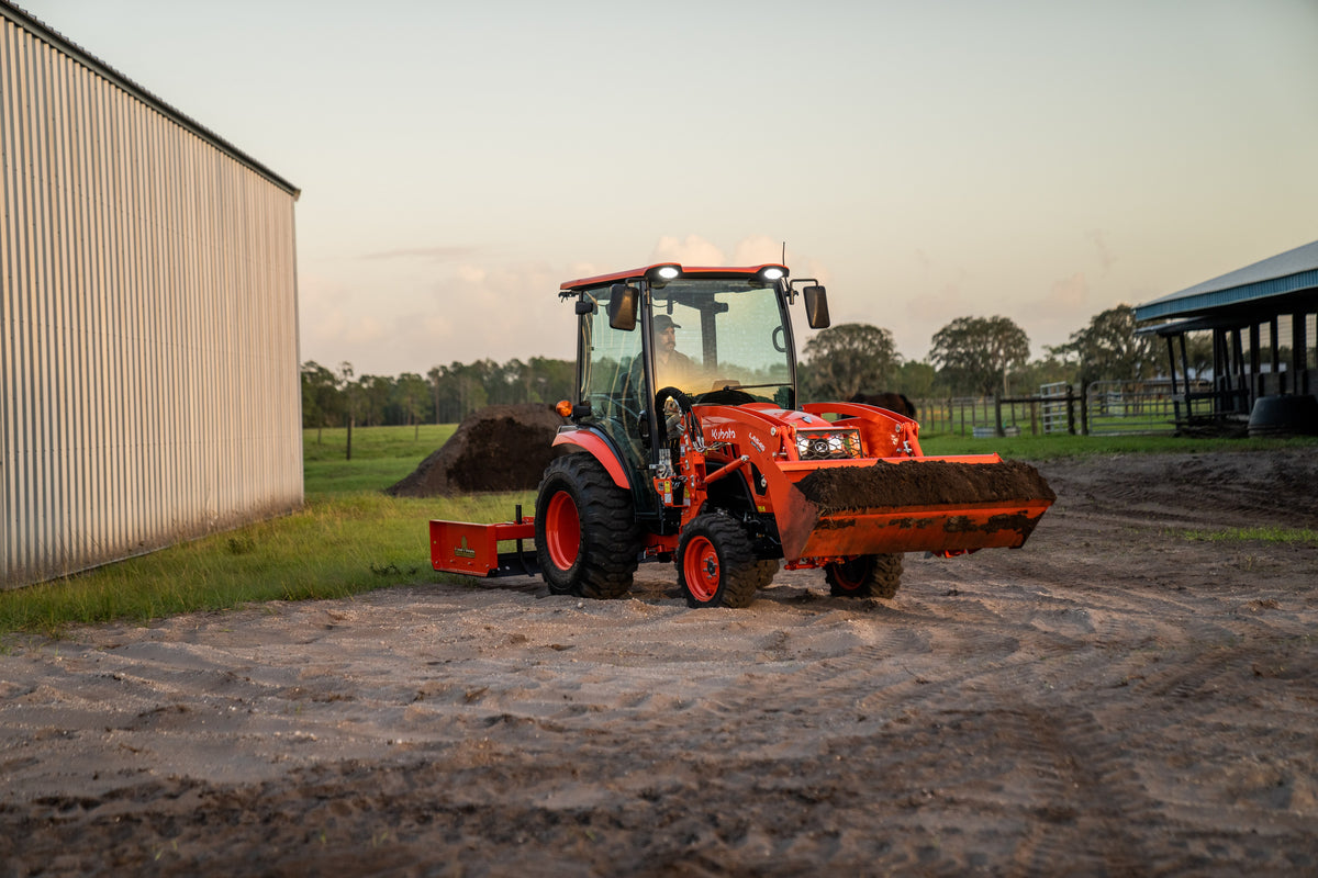 Kubota LX4020 cab tractor with a full front bucket and a rear box blade, its work lights on beside a steel building at dusk