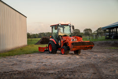 Kubota LX4020 cab tractor with a full front bucket and a rear box blade, its work lights on beside a steel building at dusk