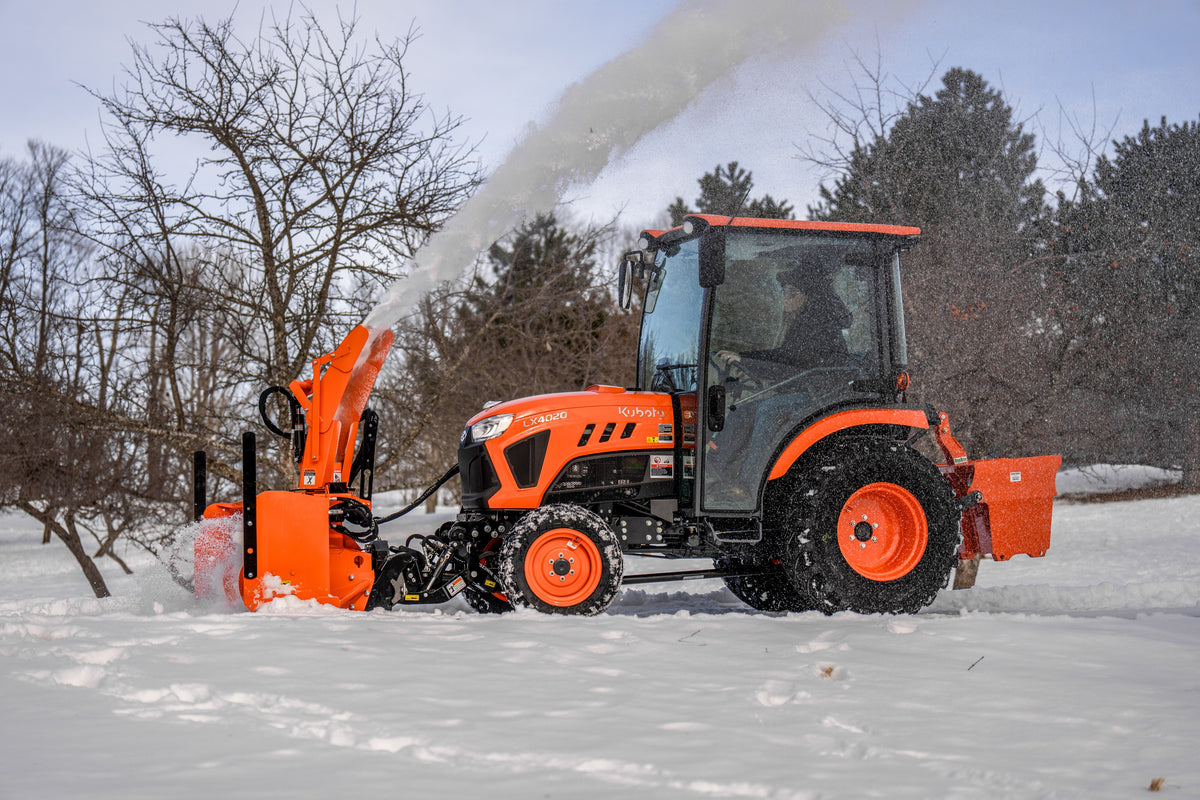 Kubota LX4020 cab tractor with a front snowblower and a rear ballast box, blowing snow by bare trees