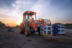Kubota M62 tractor-loader-backhoe lifting a loaded pallet with pallet forks on a building site at sunset