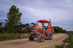 Kubota M62 tractor-loader-backhoe carrying brush in a loader grapple along a gravel road