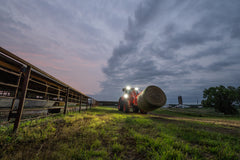 Kubota M7 series cab tractor carrying a round bale on its front loader past livestock pens at dusk
