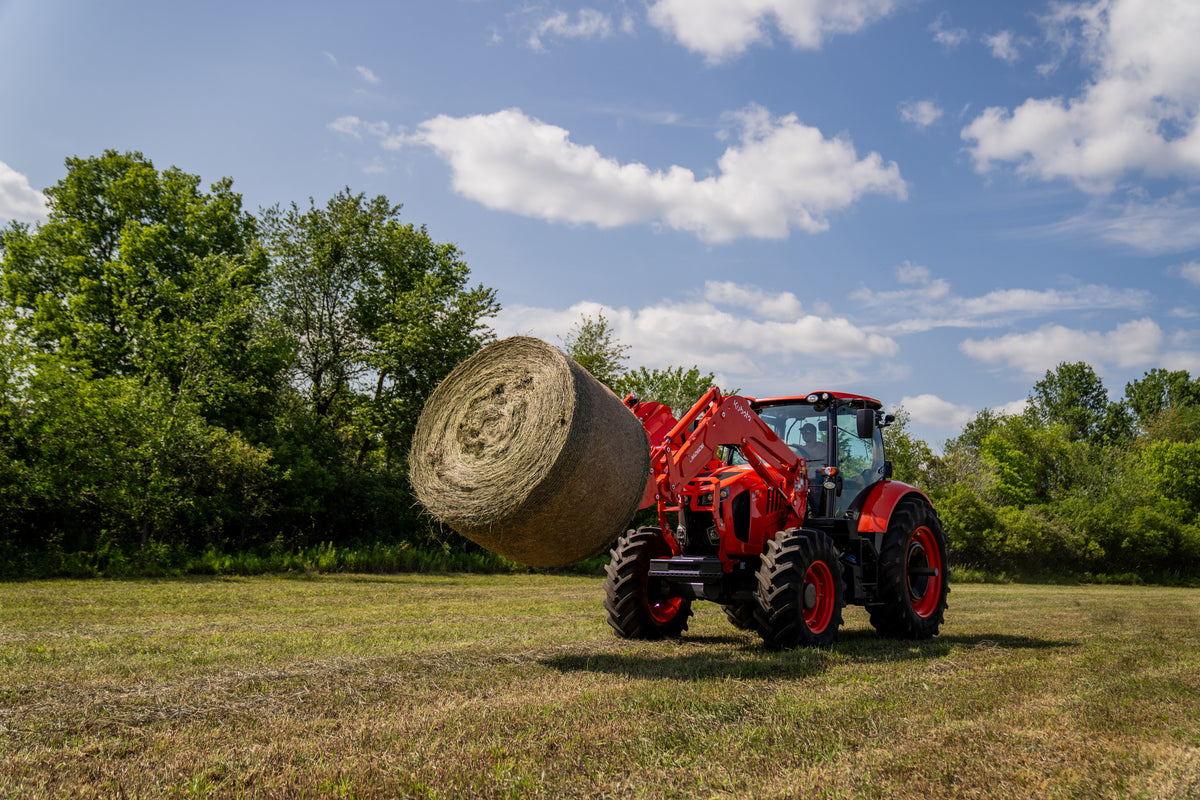 Kubota M7 series cab tractor carrying a round bale on its front loader across a hay field