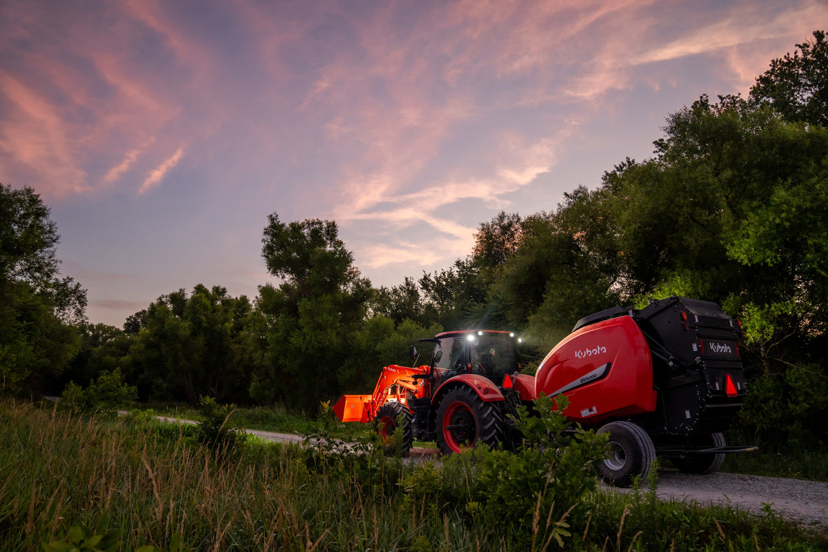 Kubota M7 series cab tractor with a front loader and a Kubota BV5200 round baler at dusk