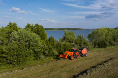 Kubota M7 series cab tractor with a front loader, pulling a round baler through a hay field beside a lake