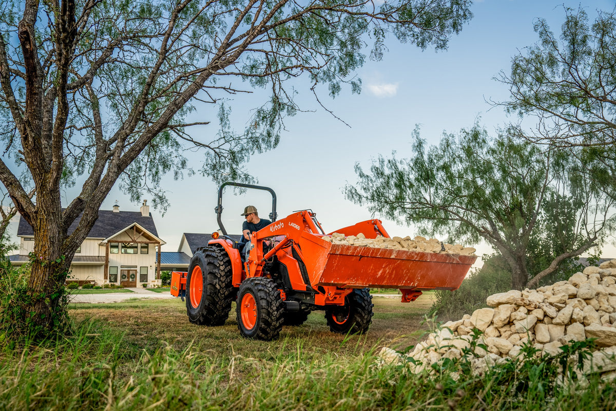 Kubota MX4900 carrying a loader bucket of rock past a farmhouse
