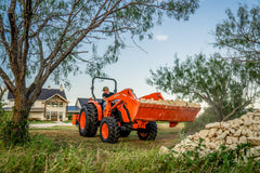 Kubota MX4900 carrying a loader bucket of rock past a farmhouse