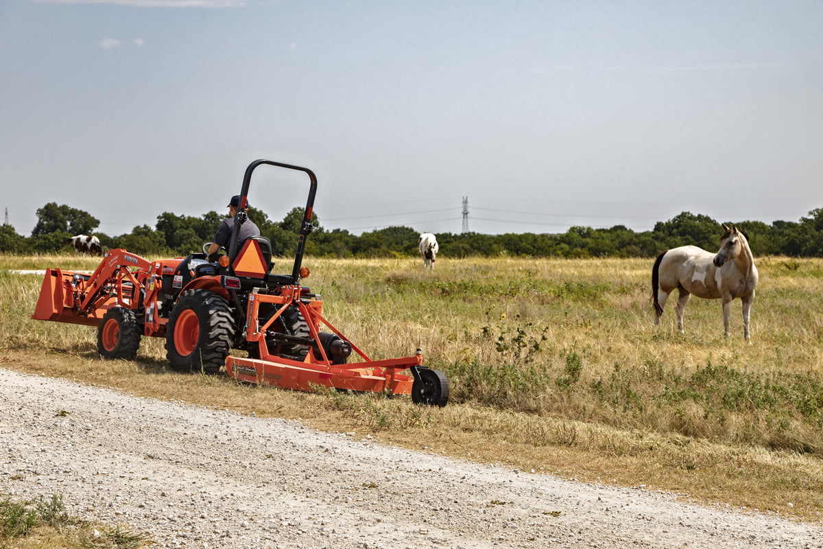 A man mowing a field edge beside a gravel road with a Land Pride RCR1248 rotary cutter behind a Kubota compact tractor with a front loader, horses grazing behind