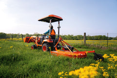 A woman mowing tall grass and wildflowers by a wire fence with a Land Pride RCR12 series rotary cutter behind a Kubota sub-compact tractor with a canopy and front loader