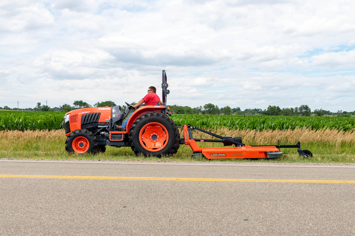 A man mowing a roadside with a Land Pride RCR1884 rotary cutter behind a Kubota L5060 tractor, a cornfield behind