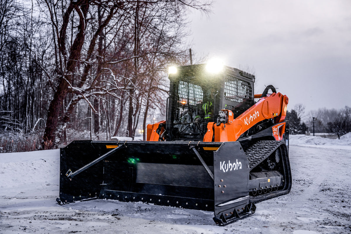 Kubota compact track loader with its lights on and a Kubota snow pusher, in snow at dusk