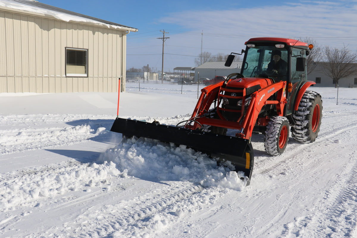 Kubota tractor with a cab and front loader pushing snow with a Land Pride STB15 series snow trip blade beside a steel building