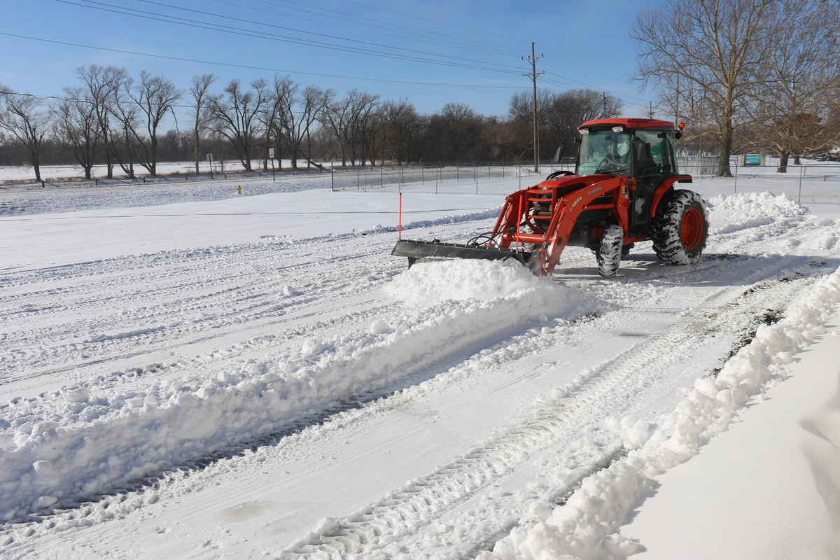 Kubota tractor with a cab and front loader clearing a snowy lot with a Land Pride STB15 series snow trip blade