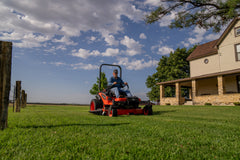 Kubota ZD1211 zero-turn mower mowing a lawn by a fence line, a farmhouse behind