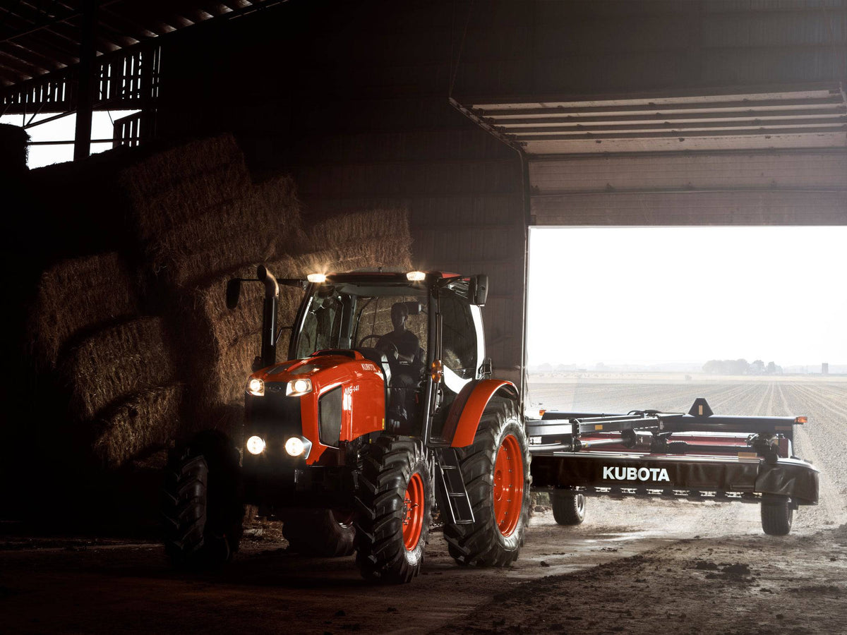 Kubota M6 series cab tractor pulling a Kubota disc mower out of a barn, its lights on