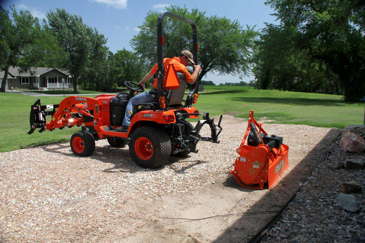 A man hitching a Land Pride RTR05 series rotary tiller to a Kubota sub-compact tractor on a gravel pad