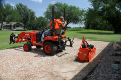 A man hitching a Land Pride RTR05 series rotary tiller to a Kubota sub-compact tractor on a gravel pad