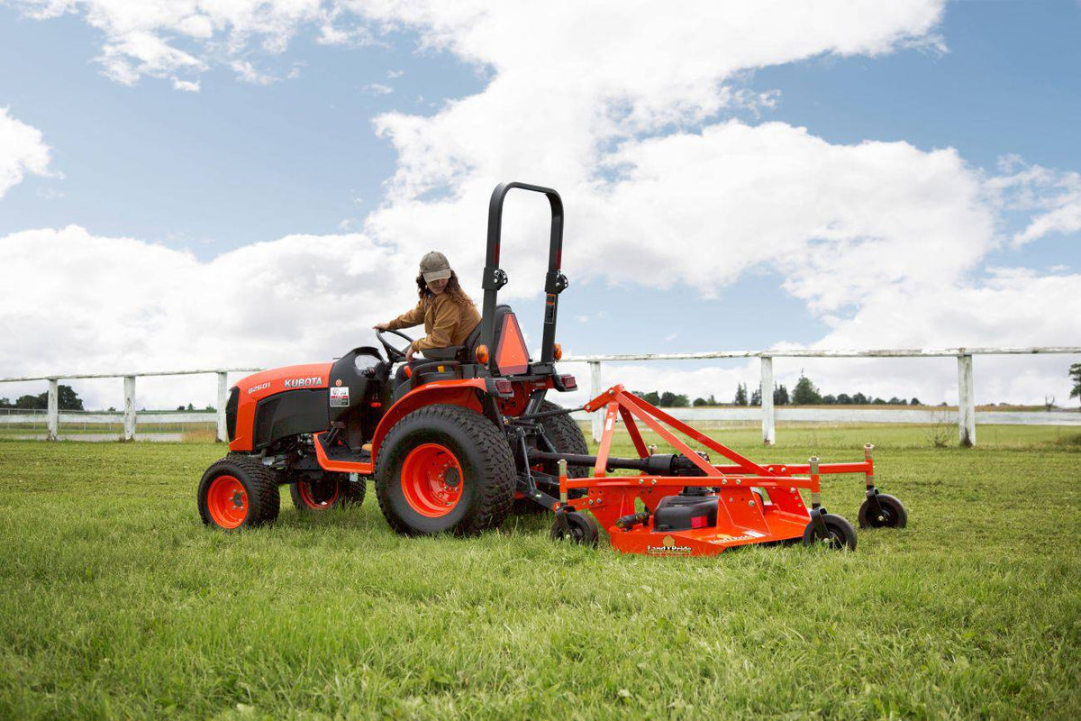 A woman mowing a pasture with a Land Pride FDR16 series grooming mower behind a Kubota compact tractor, a white fence behind