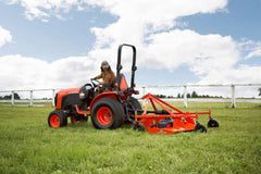 A woman mowing a pasture with a Land Pride FDR16 series grooming mower behind a Kubota compact tractor, a white fence behind