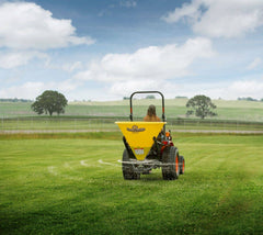A woman spreading across a pasture with a Land Pride FSP series broadcast spreader on a Kubota compact tractor, seen from behind