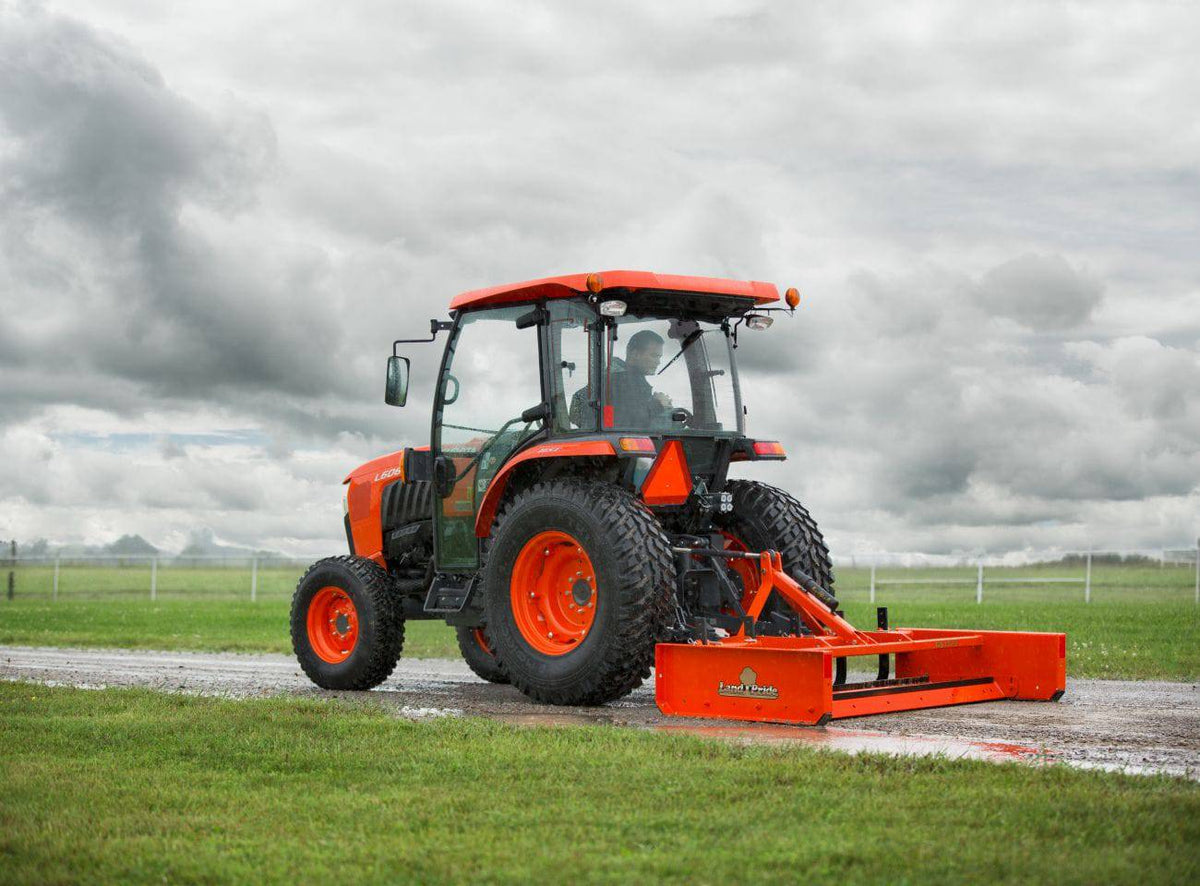 Kubota tractor with a cab pulling a Land Pride GS15 series grading scraper along a gravel lane
