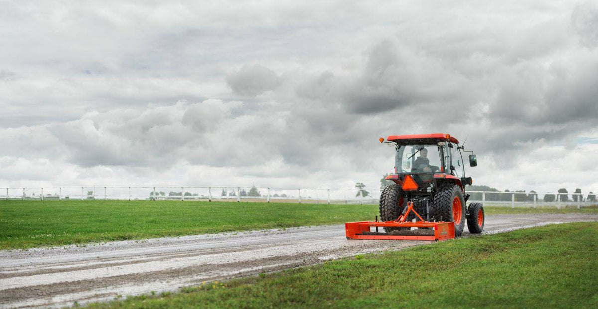 Kubota tractor with a cab grading a gravel lane with a Land Pride GS15 series grading scraper, under a cloudy sky