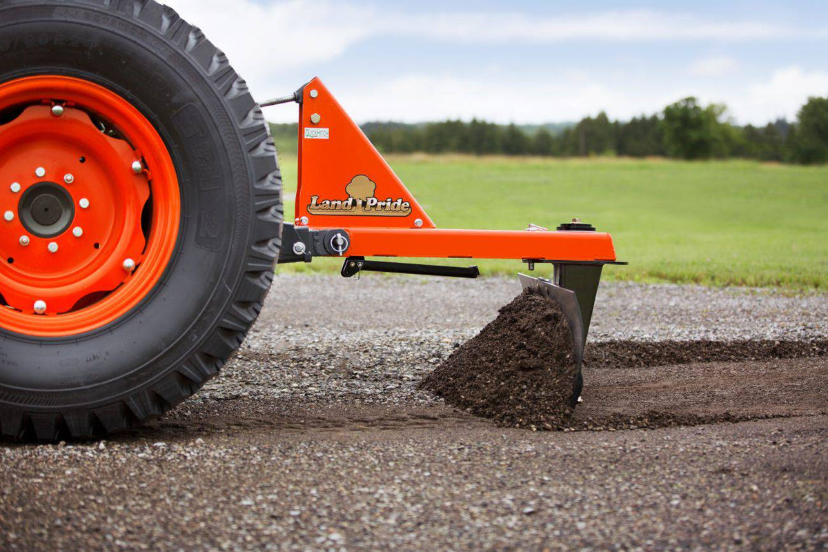 A Land Pride RB16 series rear blade pushing dirt across a gravel drive, beside a Kubota tractor's rear tire