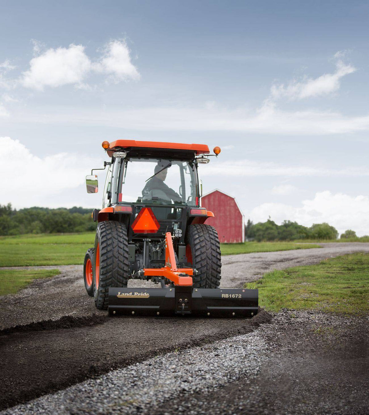 A Kubota tractor with a cab grading a gravel lane with a Land Pride RB1672 rear blade, a red barn behind