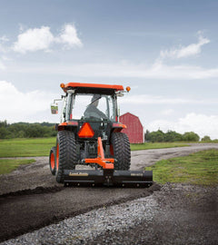 A Kubota tractor with a cab grading a gravel lane with a Land Pride RB1672 rear blade, a red barn behind