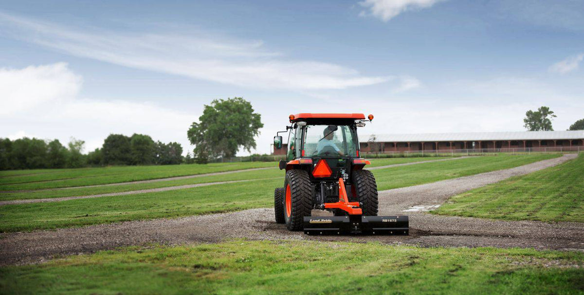 A Kubota tractor with a cab using a Land Pride RB1672 rear blade on a gravel lane between mown fields