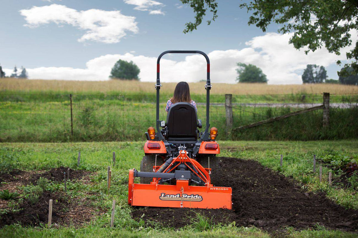 A woman tilling a garden with a Land Pride RTR05 series rotary tiller on a Kubota sub-compact tractor, rear view