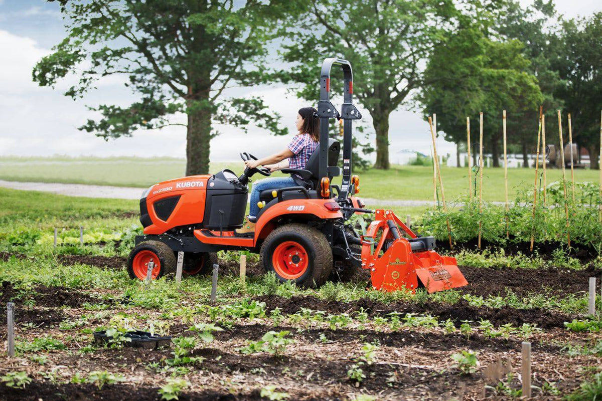 A woman driving a Kubota sub-compact tractor with a Land Pride RTR05 series rotary tiller along a garden row