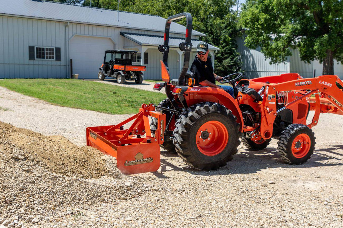 A man smoothing gravel with a Land Pride BB12 series box scraper on a Kubota compact tractor with a front loader, a utility vehicle parked behind