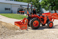 A man smoothing gravel with a Land Pride BB12 series box scraper on a Kubota compact tractor with a front loader, a utility vehicle parked behind