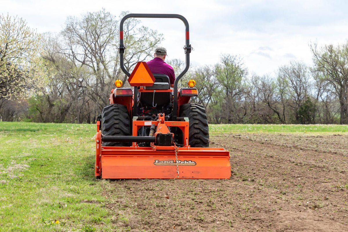 A man tilling a field with a Land Pride RTR12 series rotary tiller on a Kubota compact tractor, rear view
