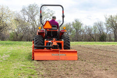 A man tilling a field with a Land Pride RTR12 series rotary tiller on a Kubota compact tractor, rear view