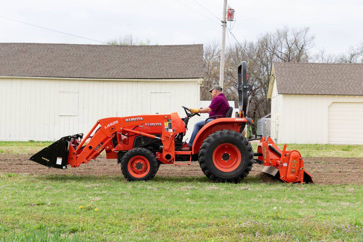 A Kubota compact tractor with a front loader tilling beside white buildings with a Land Pride RTR12 series rotary tiller