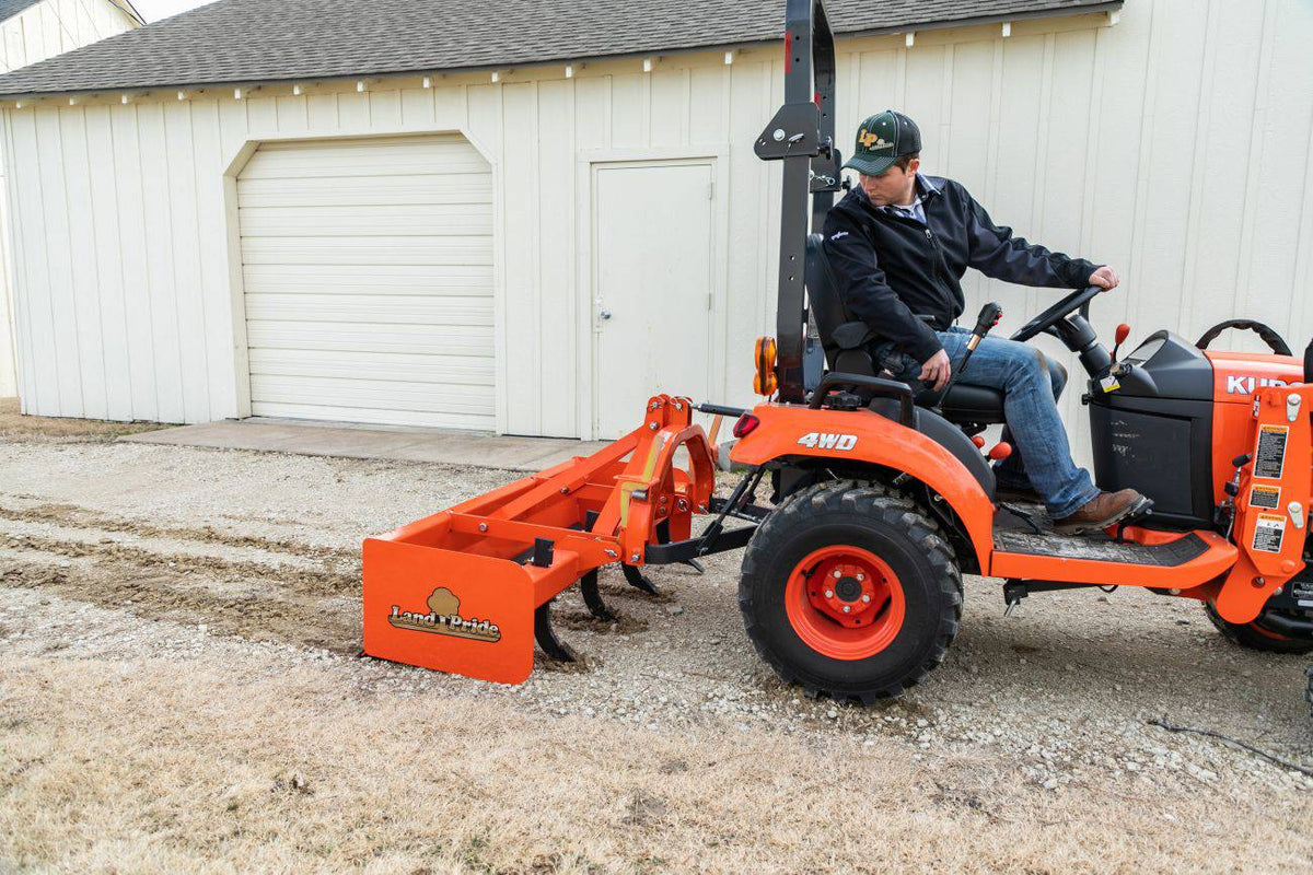 A man scraping a gravel yard with a Land Pride BB12 series box scraper behind a Kubota sub-compact tractor, beside a white barn