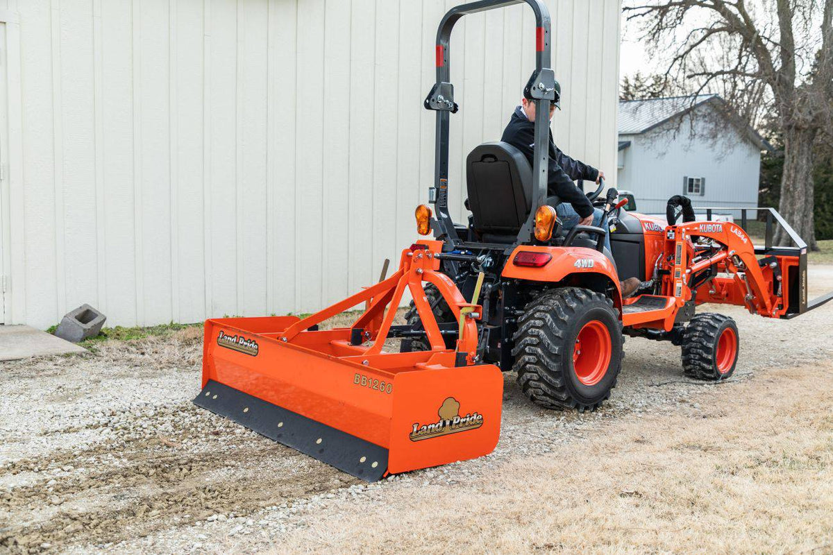 A Land Pride BB1260 box scraper on a Kubota sub-compact tractor with a front loader and pallet forks, beside a white barn