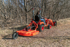A woman on a Kubota sub-compact tractor with a front loader, cutting brush with a Land Pride RCR12 series rotary cutter, rear view