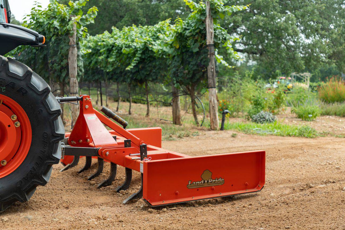 A Land Pride GS25 series grading scraper with its scarifier shanks down, behind a Kubota tractor in a vineyard