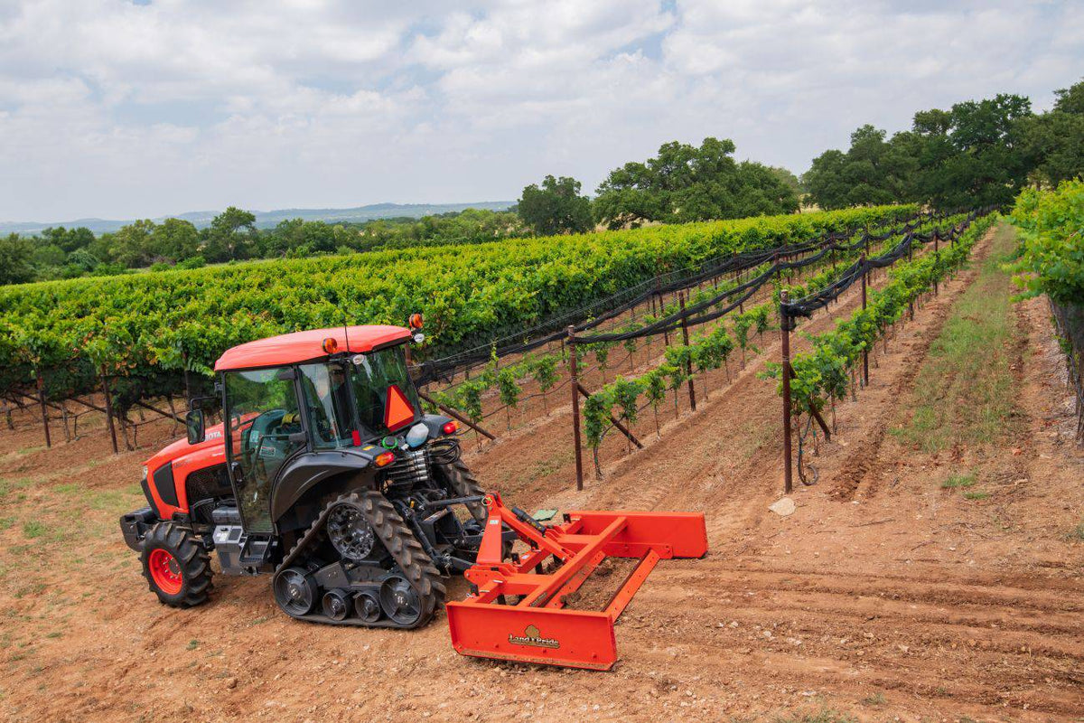 A Kubota tractor on rubber tracks pulling a Land Pride GS25 series grading scraper between vineyard rows